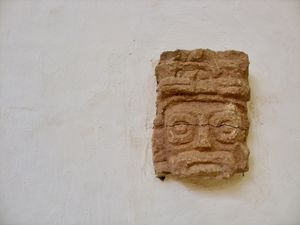Close-up of a small Mayan stone carving of a face, exhibited by itself on a white plaster museum wall.
