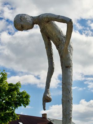 A very tall, skinny sculpture of a human leans over and is seen pointing down at the ground.