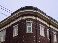 A symmetrical view looking up at the rounded corner of an old mid-rise dark brown brick building with beigh trim. Power lines cross the frame at a diagonal angle in front of the building.