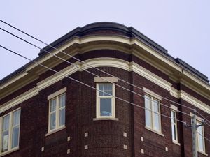 A symmetrical view looking up at the rounded corner of an old mid-rise dark brown brick building with beigh trim. Power lines cross the frame at a diagonal angle in front of the building.