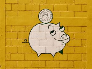 An orthogonal view of a birght yellow block wall with the logo in white painted on it. The logo is a smiling piggy bank with a 50-cent coin dropping into the slot on its back.