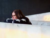 A distant candid portrait of a beautiful woman wearing glasses and resting her head on a wall as she stares at something out of frame.