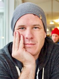 Head and shoulders portrait of a man in a hoodie and winter hat. His chin is resting in his hands and he has a serious look on his face as he stares directly into the camera.