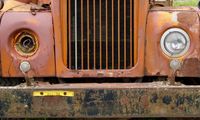 Close-up view of an abandoned truck's grill, bumper, and headlights. It's a nice shade of rusty orange, severley weathered, missing a headlight, and part of the license plate has been torn off.