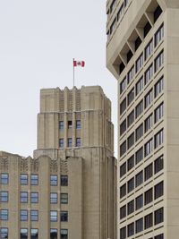 An angled view of a couple of beige government buildings—one older and the other a bit more modern. The older building is striking in an art deco style with a Canadian flag flying from its rooftop.