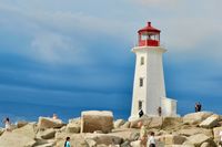The famous lighthouse towers above scattered rocks and tourists in the foreground, agasint the backdrop of a deep blue overcast sky.
