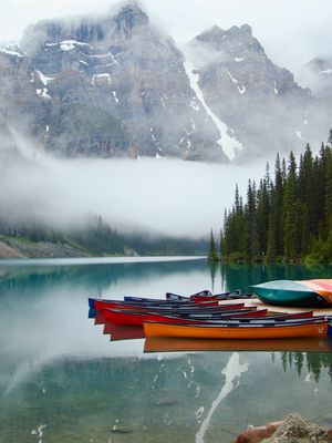 In the foreground more than a dozen canoes in different colors sit empty at a dock while the snowy mountain peaks stand tall above Moraine Lake's crystal clear water and a cloud bank passes through.