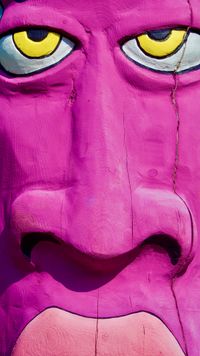 A very tall close-up of a wooden sculpture of a stylized face. The face is painted bright fuchsia with yellow eyes and pink lips.