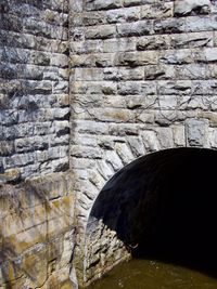 Water flows slowly out of an arched tunnel build into a rough stone wall covered in vines.