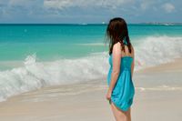 Side profile of a woman in a blue sundress looking away from the camera at the waves crashing on a sunny tropical beach.