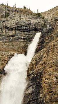 Looking up at the canyon as it opens up to a black and brown cliff face and white water pours out and tumbles down.