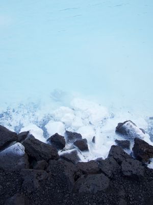 Looking down at rough lava rocks against the blue/turquoise water of a hot spring. Where the water and rocks meet it's foamy and bubbly.