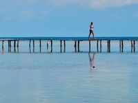 Candid scene of a woman walking along a boardwalk raise above the water with nothing behind her but the horizon and the sky. She is partially reflected in the calm water in the foreground.