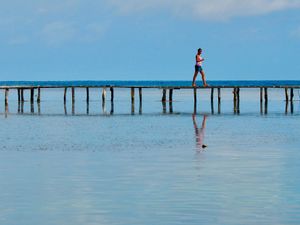 Candid scene of a woman walking along a boardwalk raise above the water with nothing behind her but the horizon and the sky. She is partially reflected in the calm water in the foreground.
