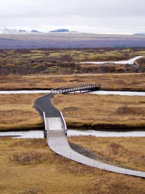 A pathway and some bridges meander into the distance in this landscape shot. Lava fields stretch out to the horizon where snow-covered mountains can be seen.