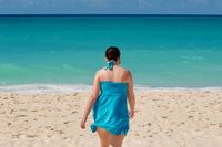 Candid shot of a woman in a blue sundress walking away from the camera towards the very blue tropical ocean. The wind is blowing her dress to the side.