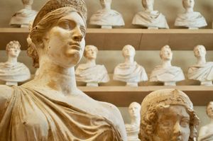 In the foreground is the head and shoulders of a statue of a woman. In the background are three shelves of stones busts of various Roman men.