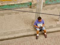 Candid street scene looking down at a man sitting on the sidewalk holding an FM radio.