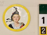An abstract of a wall at a curling rink, featuring part of a scoreboard and a framed vintage painting of a woman in Scottish dress.