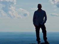 The silhouette of a man in hiking clothes standing on a rock on a mountain peak with nothing but open sky and foothills behind him.