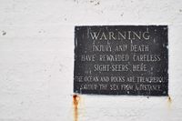 Close-up of a black plaque on a worn white wall that reads, 'WARNING. INJURY AND DEATH HAVE REWARDED CARELESS SIGHT-SEERS HERE. THE OCEAN AND ROCKS ARE TREACHEROUS. SAVOUR THE SEA FROM A DISTANCE.'