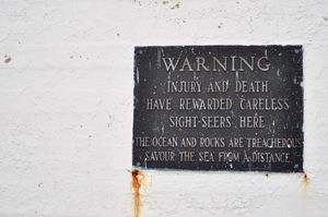 Close-up of a black plaque on a worn white wall that reads, 'WARNING. INJURY AND DEATH HAVE REWARDED CARELESS SIGHT-SEERS HERE. THE OCEAN AND ROCKS ARE TREACHEROUS. SAVOUR THE SEA FROM A DISTANCE.'