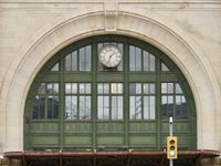 Street level view of the green archway and windows above the main entrance to Winnipeg's train station. The analog clock shows 1:34pm in roman numerals.