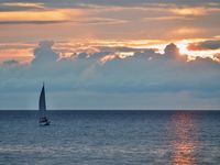 A sailboat heads towards the horizon as the sun sets behind a large cloud bank.