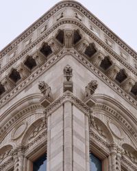 Looking up at the corner of an intricately decorated grey building against a grey sky. Three lion statues are perched against the wall looking down.