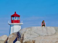The top of a lighthouse stands above large granite boulders in the foreground. A woman is sitting on top of the boulder with her back to the camera.