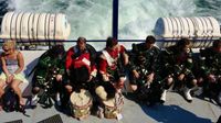 A candid shot of bagpipers and drummers in full dress taking a break while riding on a ferry. They are sitting in a row chatting with the wake of the ferry boat trailing behind them.