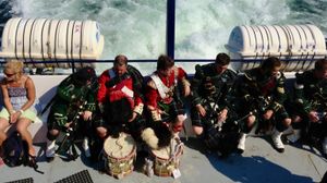 A candid shot of bagpipers and drummers in full dress taking a break while riding on a ferry. They are sitting in a row chatting with the wake of the ferry boat trailing behind them.