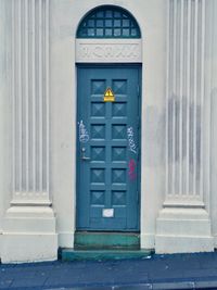 An orthogonal view of the simple building with a green door center frame. The door has an electricaly warning symbole on it and some graffiti scribbled on it.