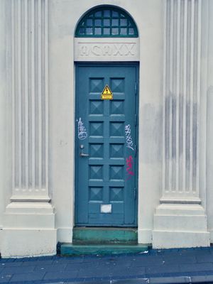An orthogonal view of the simple building with a green door center frame. The door has an electricaly warning symbole on it and some graffiti scribbled on it.