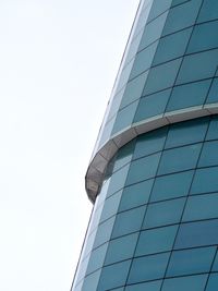 Looking up at two levels of blue-green window surfaces arcing away from the camera against a grey sky.