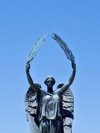 A statue of an angelic figure holding olive branches over their head is set against a bright blue sky.