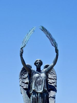 A statue of an angelic figure holding olive branches over their head is set against a bright blue sky.