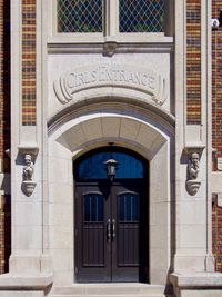 An orthogonal view of an old school entrance. The door is framed by gargoyle sculptures and an engraved sign above it that reads GIRLS' ENTRANCE.