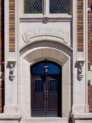 An orthogonal view of an old school entrance. The door is framed by gargoyle sculptures and an engraved sign above it that reads GIRLS' ENTRANCE.